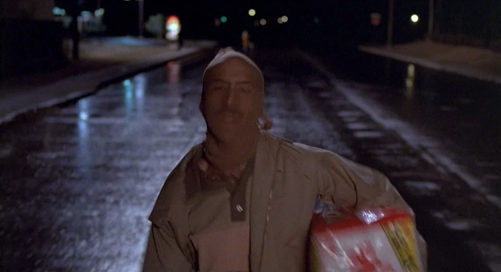 Raising Arizona (1987) movie still: medium — A wet asphalt street at night under dark, overcast conditions with distant blurred lights. A man…