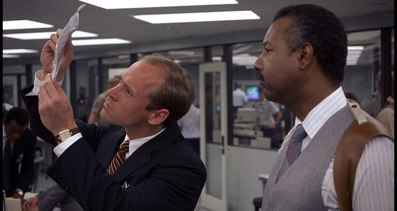 No Way Out (1987) movie still: medium — An office space with glass partitions and drop ceilings during work hours. Two men in formal business attire…