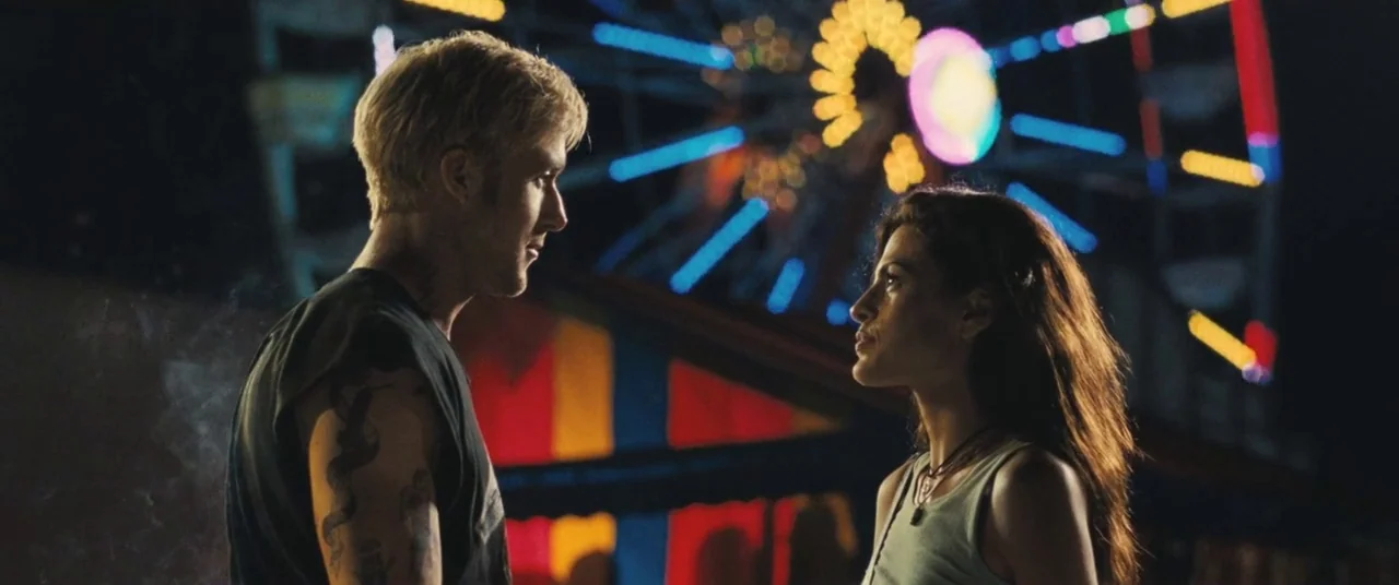 The Place Beyond The Pines (2012) movie still: medium — Two individuals stand in profile before a brightly lit, moving ferris wheel at night. The…