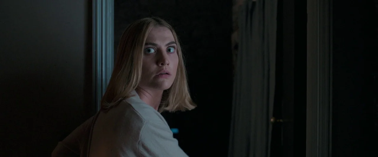 Oddity (2024) movie still: medium — A dark, shadowed space features a woman standing by a white door frame. The subject glances toward the viewer…
