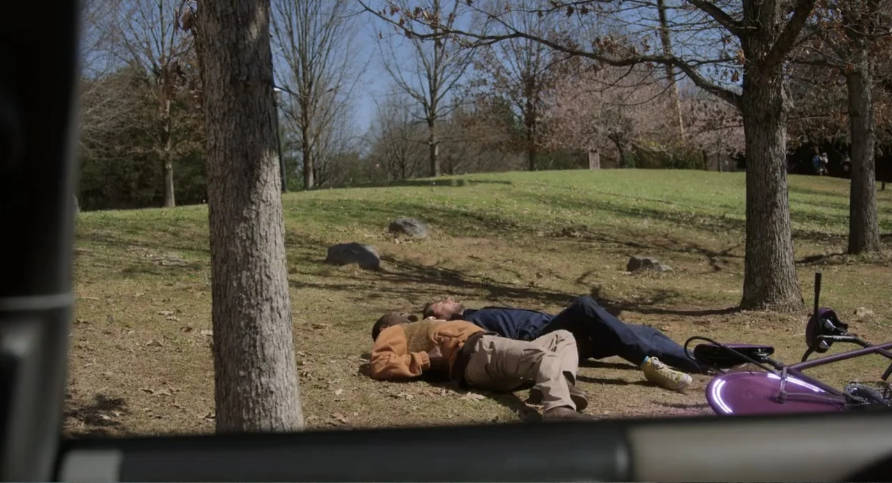 Harold and the Purple Crayon (2024) movie still: wide — A sunny public park with bare trees and a grassy slope in the background. Two men lie on the…