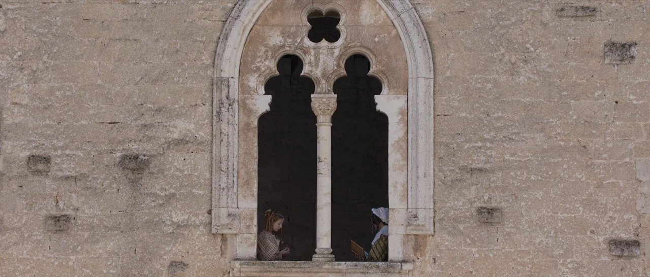 Tale of Tales (2015) movie still: wide — A stone wall features an intricate gothic arched window divided by a central column. Two women sit within…