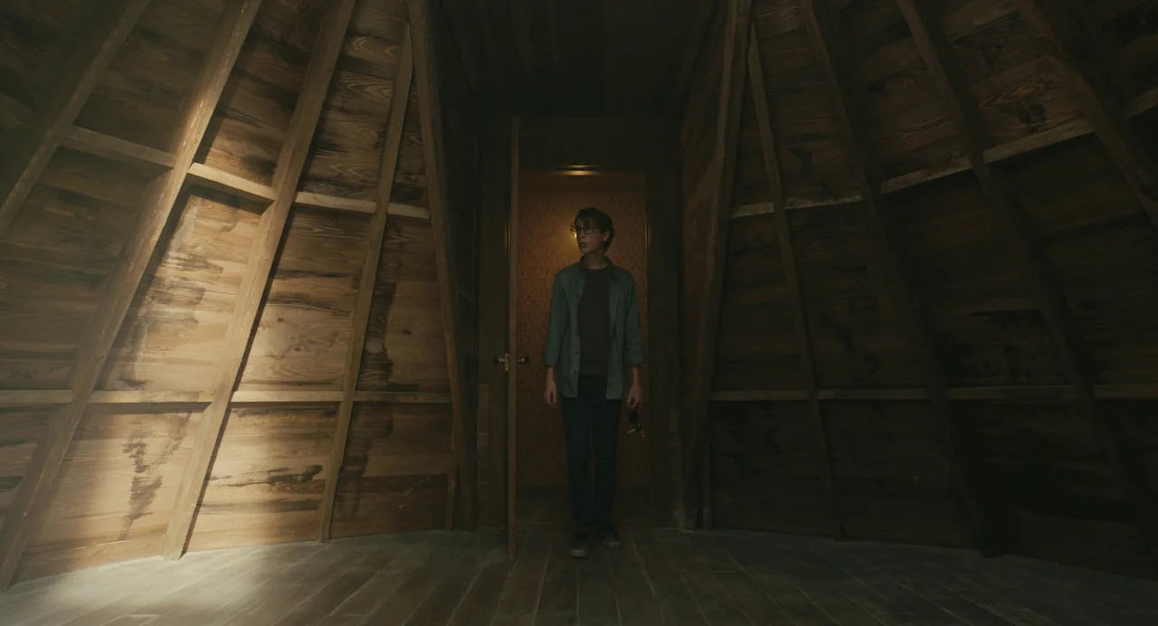 The Life of Chuck (2024) movie still: fullbody — A wooden attic features diagonal beams and a central doorway leading to another room. A teenage boy…