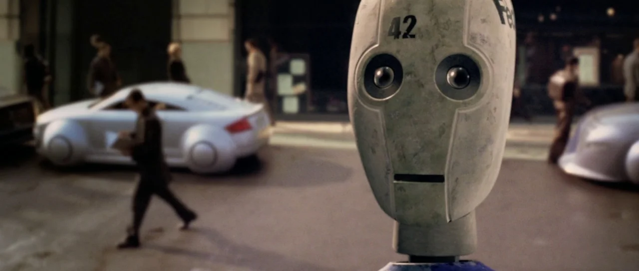 I, Robot (2004) movie still: closeup — A robotic head with the number 42 sits in the foreground of a city. Pedestrians walk past futuristic cars on…