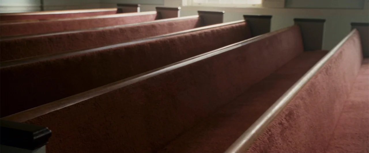 Dear John (2010) movie still: wide — Rows of wooden pews with red fabric upholstery sit inside a building. Empty seats face forward toward an unseen…