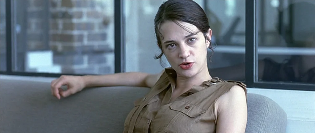 Boarding Gate (2007) movie still: medium — A woman sits on a grey sofa in a brightly lit, modern room. She looks directly forward while resting her…