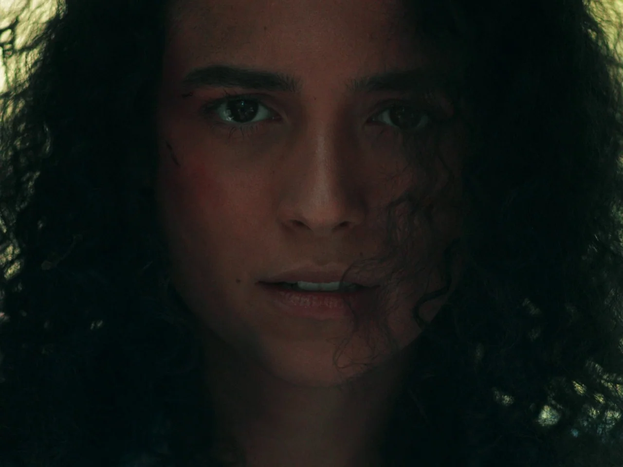 In A Violent Nature (2024) movie still: closeup — A close-up view of a person surrounded by foliage in a dim setting. The subject stares directly…