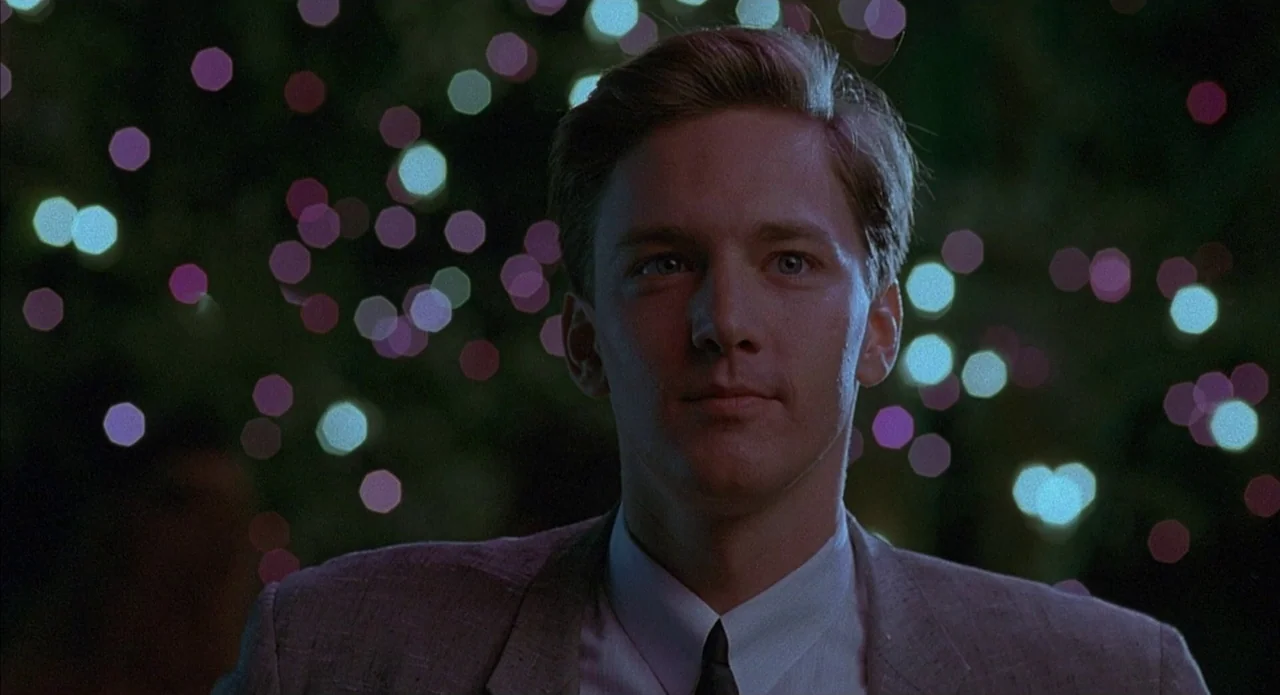 Less Than Zero (1987) movie still: medium — A man stands in an outdoor night setting with colorful circular bokeh lights behind him. The man looks…