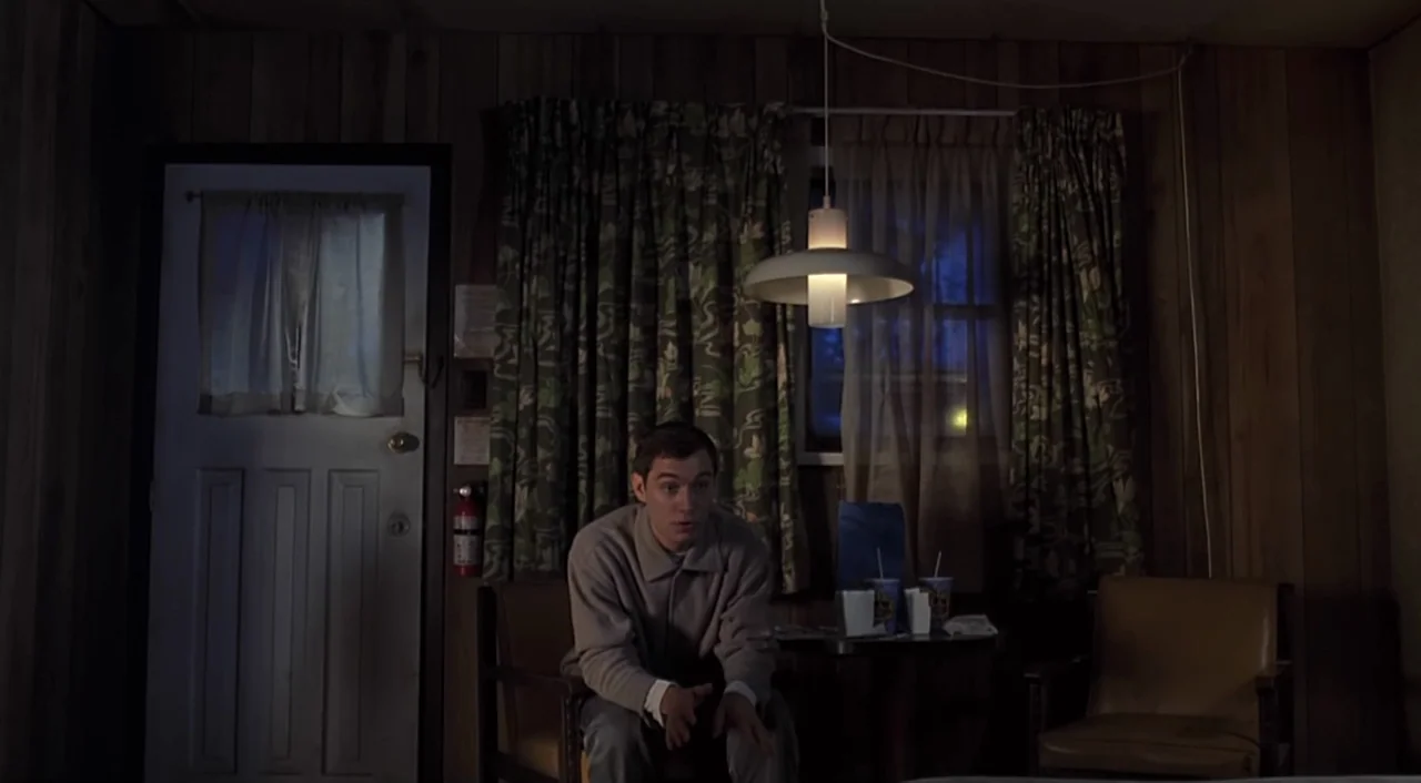 eXistenZ (1999) movie still: medium — A dimly lit motel room features brown wood-paneled walls and patterned curtains. A man sits at a small table…