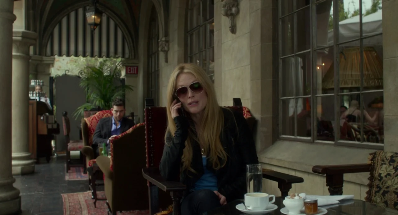 Maps to the Stars (2014) movie still: medium — An outdoor patio of a building features stone columns and a glass window facade. A blonde woman sits…