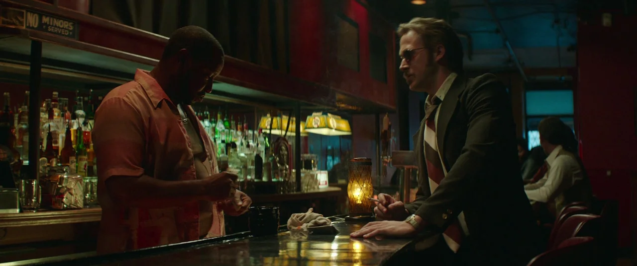 The Nice Guys (2016) movie still: medium — A dark dive bar with rows of liquor bottles and red neon signage. A man in a suit engages a bartender…
