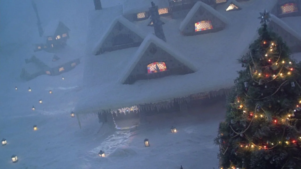 Scrooged (1988) movie still: wide — A snow-covered village features gabled houses with stained glass windows at night. A decorated Christmas tree…