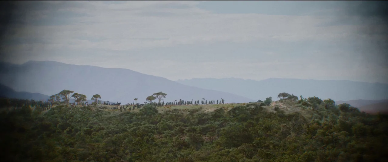 The Woman King (2022) movie still: wide — A distant ridge line shows a line of travelers moving across a grassy plain. Tall mountains rise in the…