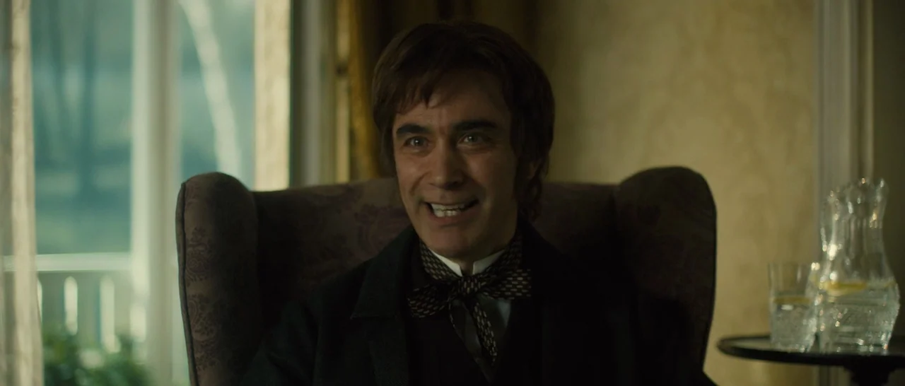 A Quiet Passion (2016) movie still: medium — A man sits in a patterned armchair in a room with yellow wallpaper. He faces the camera while smiling…