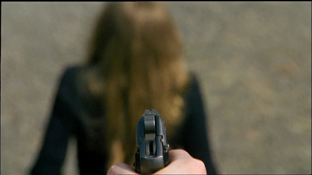 Chinese Roulette (1976) movie still: closeup — A person stands on a textured gravel surface with their back to the camera. A handgun is held in the…