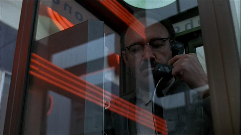The Conversation (1974) movie still: medium — A man stands inside a reflective phone booth at night. He is speaking into a telephone receiver while…