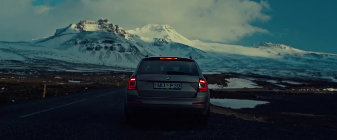 Rift (2017) movie still: wide — A silver station wagon sits on an asphalt road set against vast snowy mountains. The landscape is desolate with…