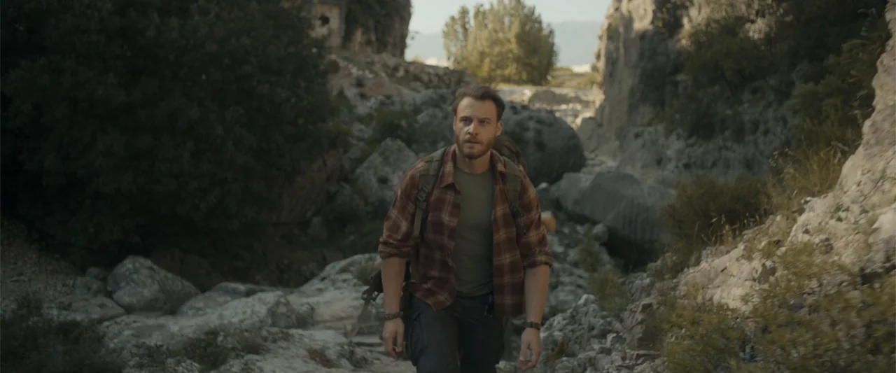 Blue Cave (2024) movie still: medium — A rocky narrow canyon path flanked by large boulders and sparse vegetation. A man wearing a backpack walks…