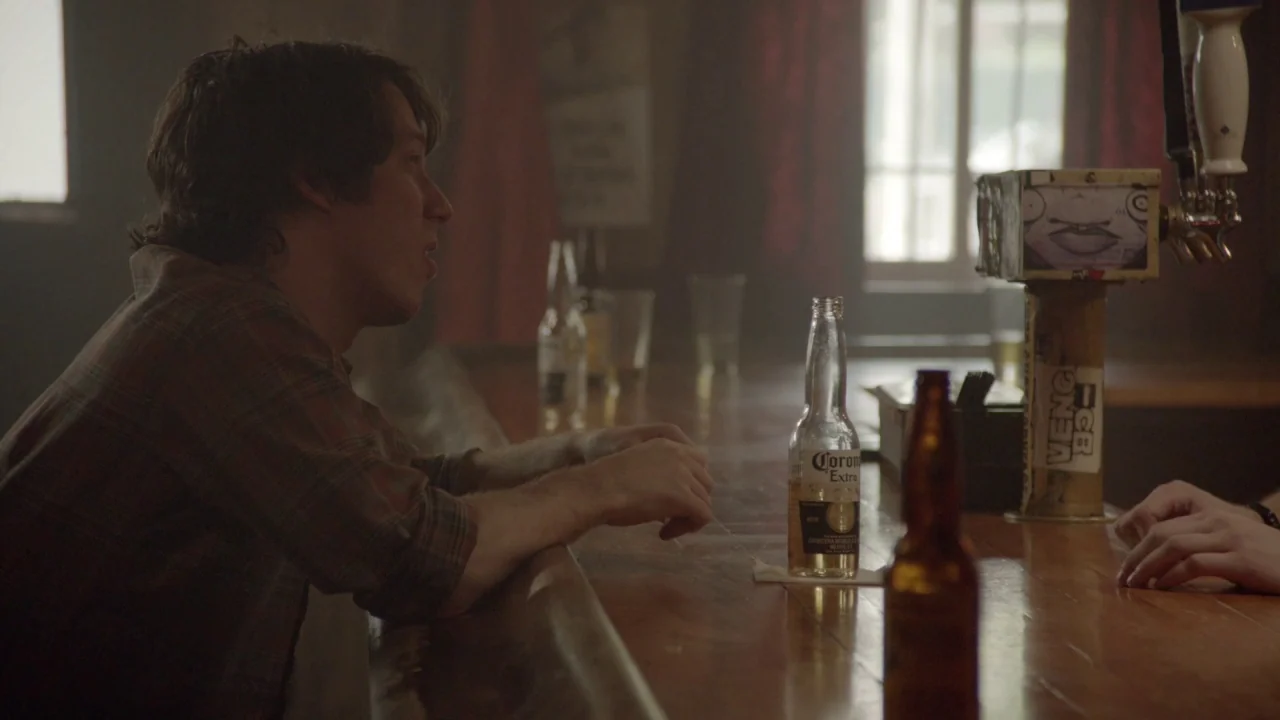 The Heart Machine (2014) movie still: medium — A dimly lit wooden bar counter features scattered glassware and a beer tap. A young man sits at the…