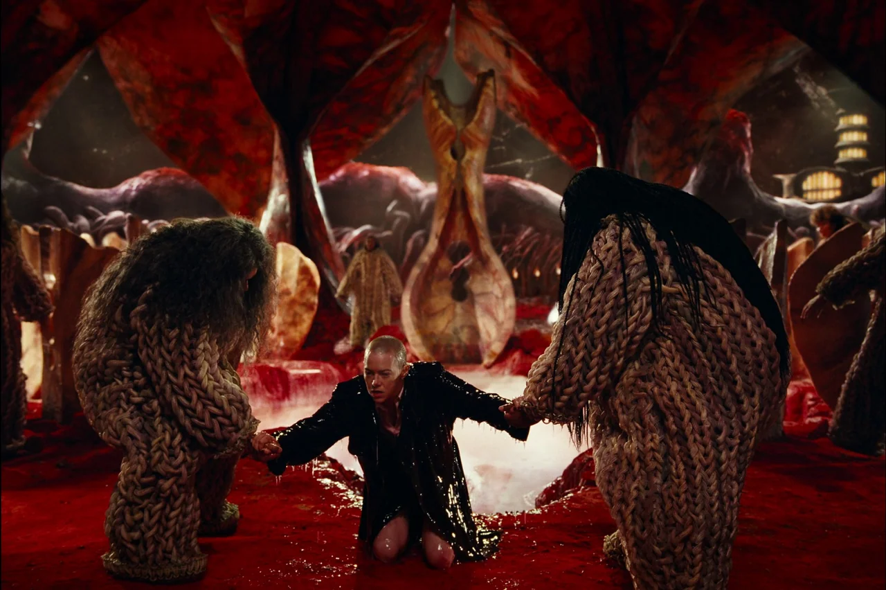 Bugonia (2025) movie still: fullbody — An organic cave structure features fleshy, vein-covered walls and a bright red floor. A bald person in a…