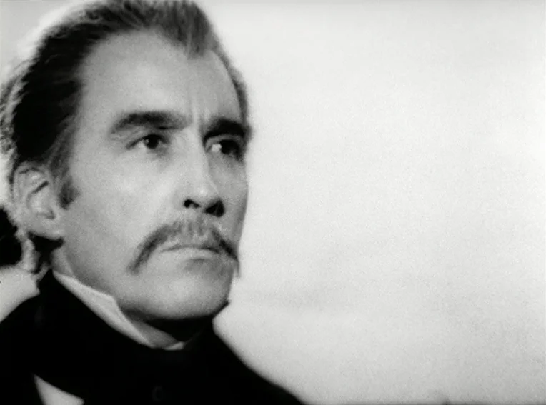 Cuadecuc, Vampir (1971) movie still: closeup — A man stands before a bright, nondescript background in a black and white film. The subject looks…