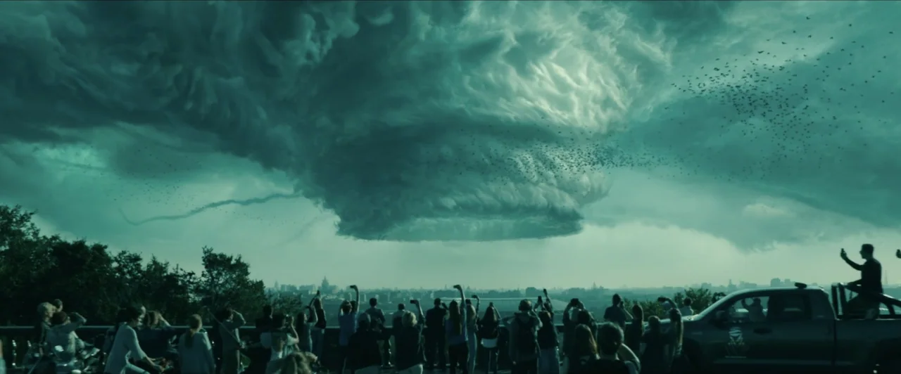 Invasion (2020) movie still: wide — An outdoor scenic overlook crowded with people looking at a massive, dark, swirling storm cloud. The group holds…