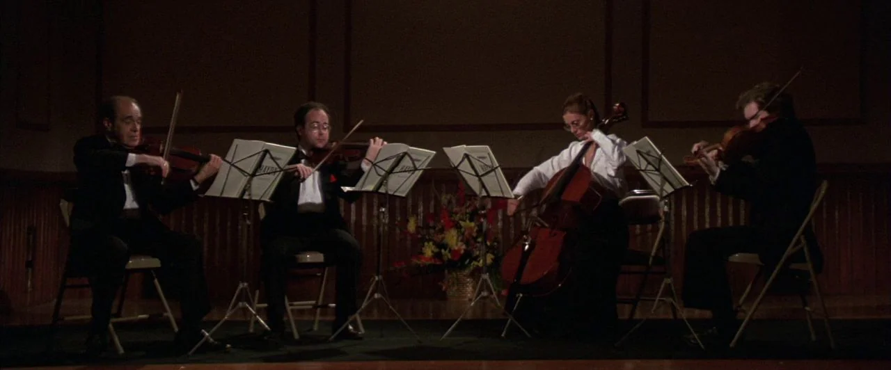 The Witches of Eastwick (1987) movie still: wide — A wooden paneled stage interior features four musicians arranged in a string quartet. The quartet…