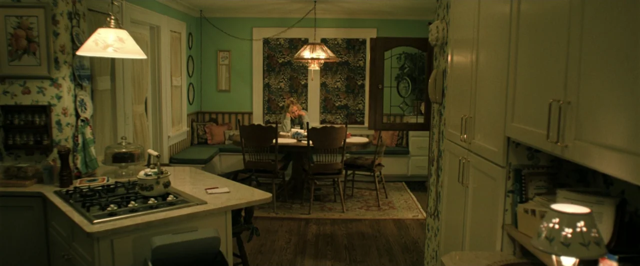 Holland (2025) movie still: wide — A green-walled kitchen features floral wallpaper and a dark wood table with chairs. A woman sits alone at the…