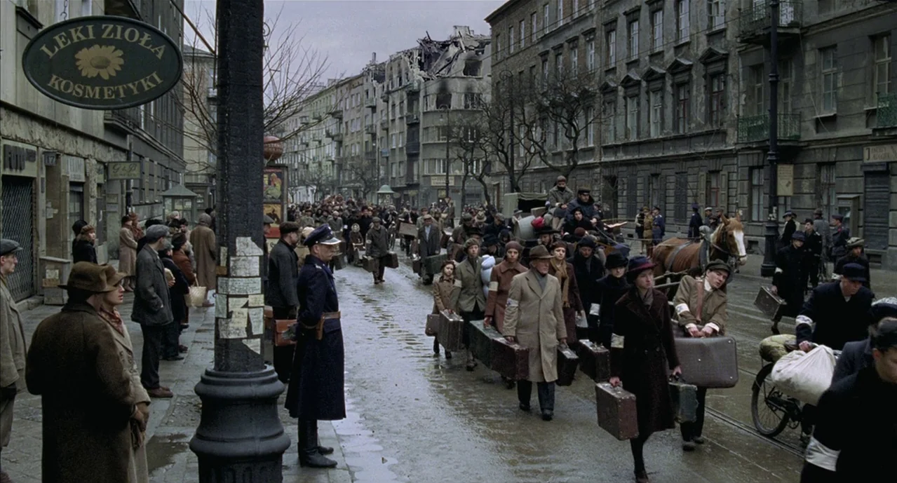 The Pianist (2002) movie still: wide — A war-torn city street in Poland features damaged buildings and a large crowd moving. Civilians carry luggage…