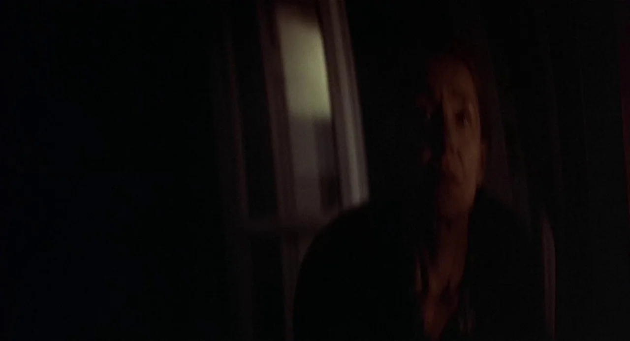 Three Colours: Blue (1993) movie still: medium — A dark, dimly lit indoor space with a single bright vertical rectangle in the background. An older…
