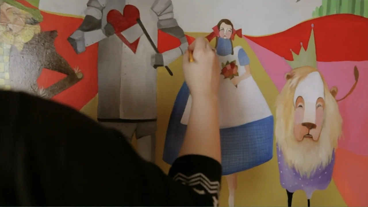 The (Dead Mothers) Club (2014) movie still: medium — An interior wall serves as a canvas for a large, flat-style painted mural. A person's hand with…