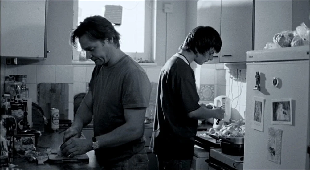 Somers Town (2008) movie still: medium — A cluttered kitchen with white-tiled walls and a refrigerator covered in photos. Two men stand facing away…