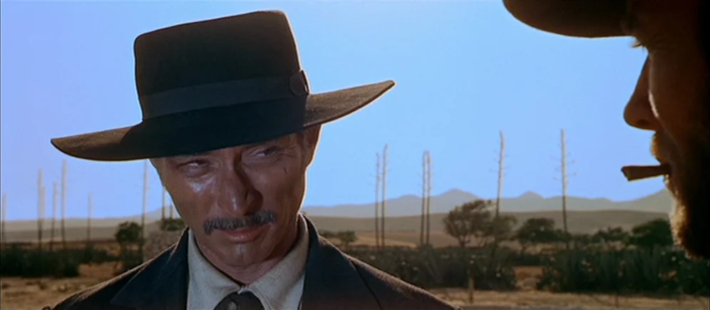 For A Few Dollars More (1965) movie still: medium — A dry, sunlit desert landscape stretches toward distant mountains under a clear blue sky. A man…
