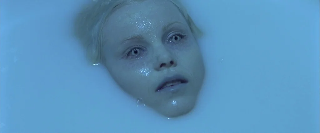The Cell (2000) movie still: closeup — A woman's face is partially submerged in a white liquid inside a bathtub. She stares upward toward the camera…