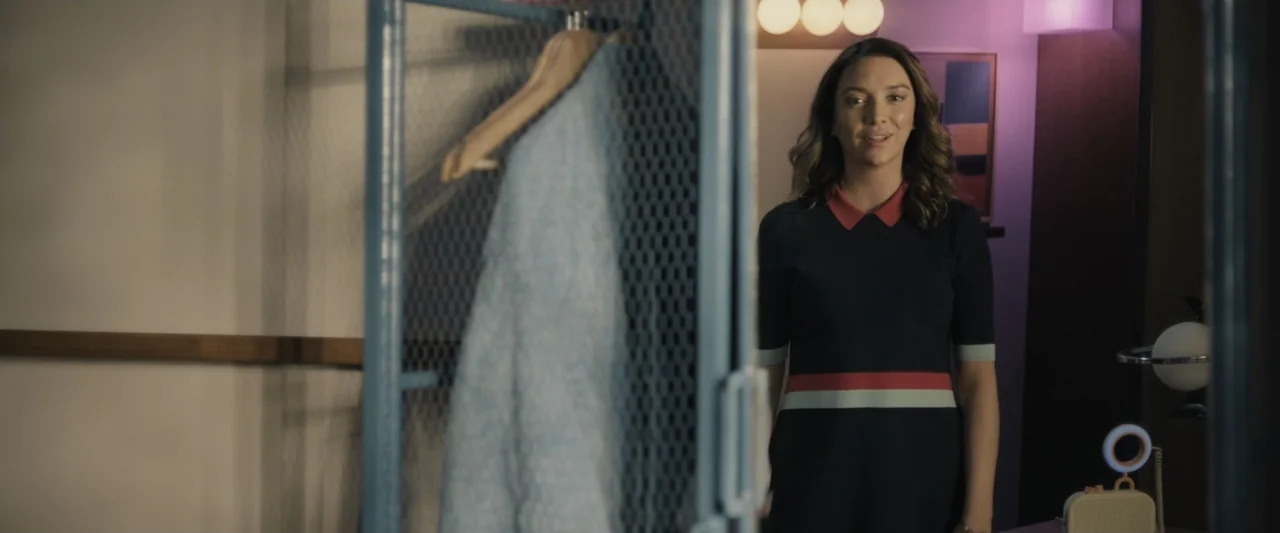 Sign Here (2017) movie still: medium — A woman stands in a modern walk-in closet with a blue wire clothing rack. She wears a navy dress and faces…
