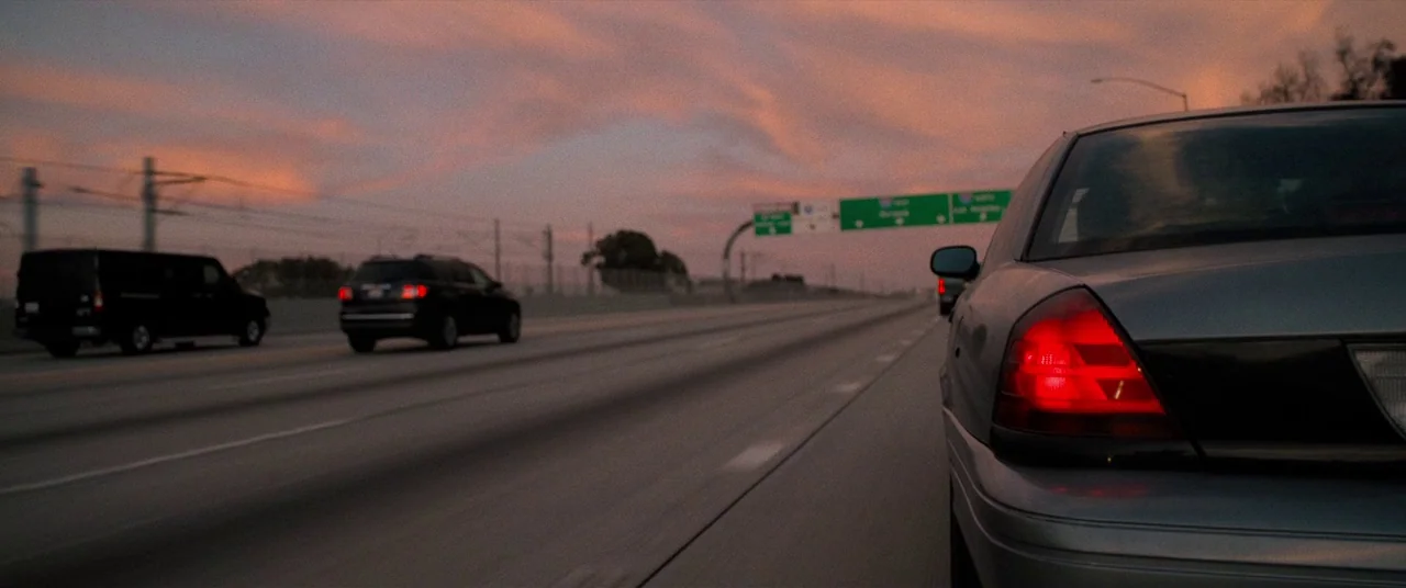 Destroyer (2018) movie still: medium — A multi-lane highway stretches into the distance under a pink and orange sunset sky. Several cars travel…