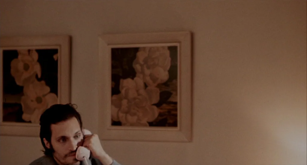 Buffalo ’66 (1998) movie still: medium — A dimly lit room features two floral art prints mounted on a cream wall. A man sits in the lower foreground…