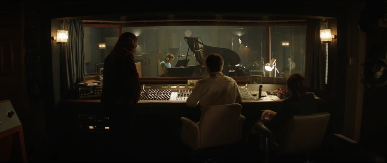 Rocketman (2019) movie still: wide — A dimly lit 1970s recording studio with a large glass partition and mixing console. Three people sit at the…