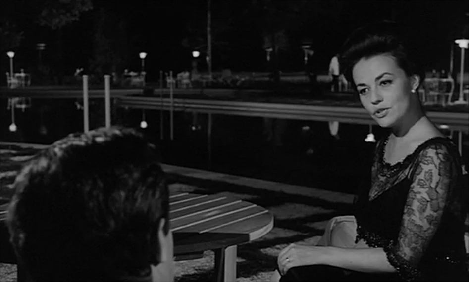 La Notte (1961) movie still: medium — A nighttime scene beside a still outdoor swimming pool with illuminated walkway lights. A woman sits across…