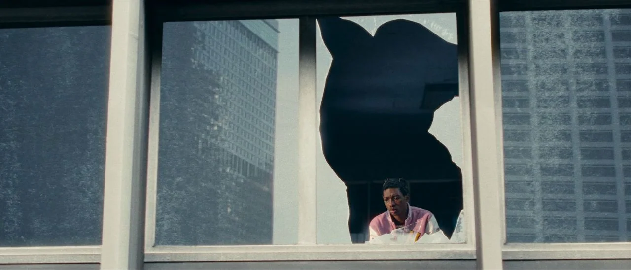 Archenemy (2020) movie still: wide — A man looks through a jagged hole in a large glass office building window. Tall buildings and sky reflect on…