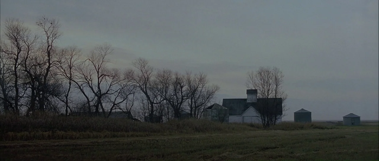 Capote (2005) movie still: establishing — A rural farmland landscape features a white barn and several metal grain silos. Bare winter trees stand in…