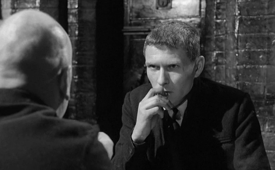 The Criminal (1960) movie still: medium — A dark, cramped space with rough stone or brick walls. A young man sits opposite an unseen person,…