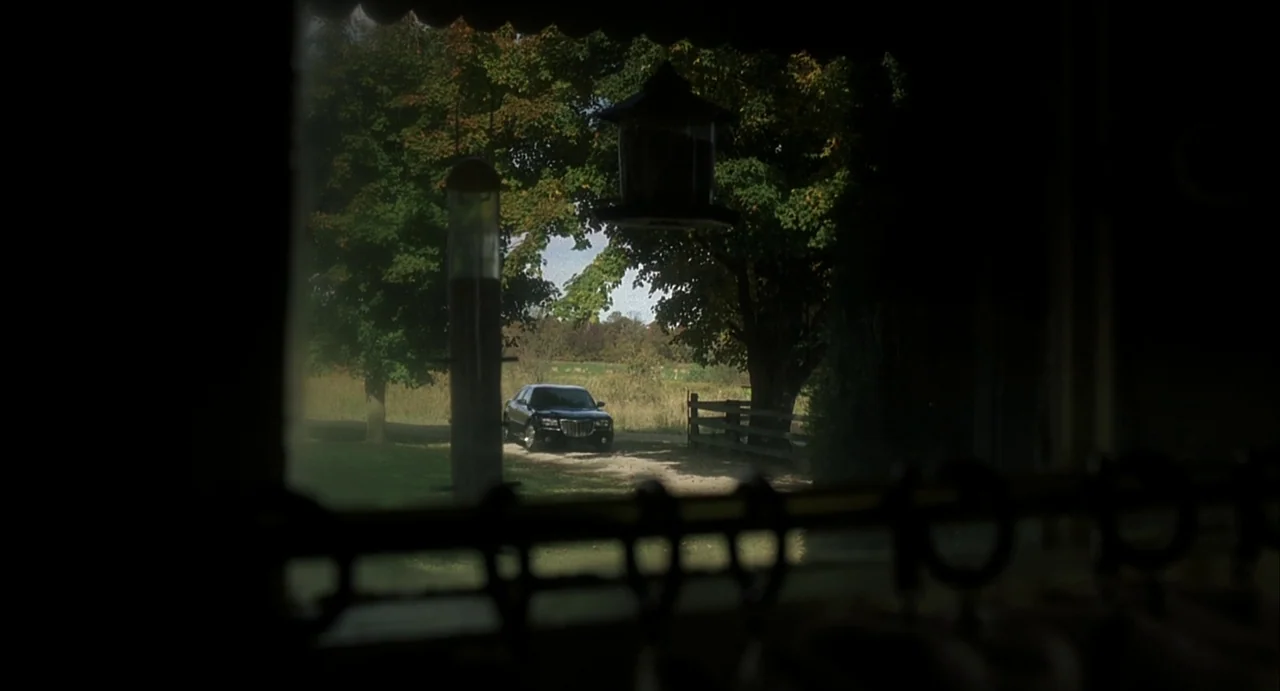 A History of Violence (2005) movie still: wide — A dark, silhouetted interior foreground looks out onto a sunlit rural property. A black sedan is…