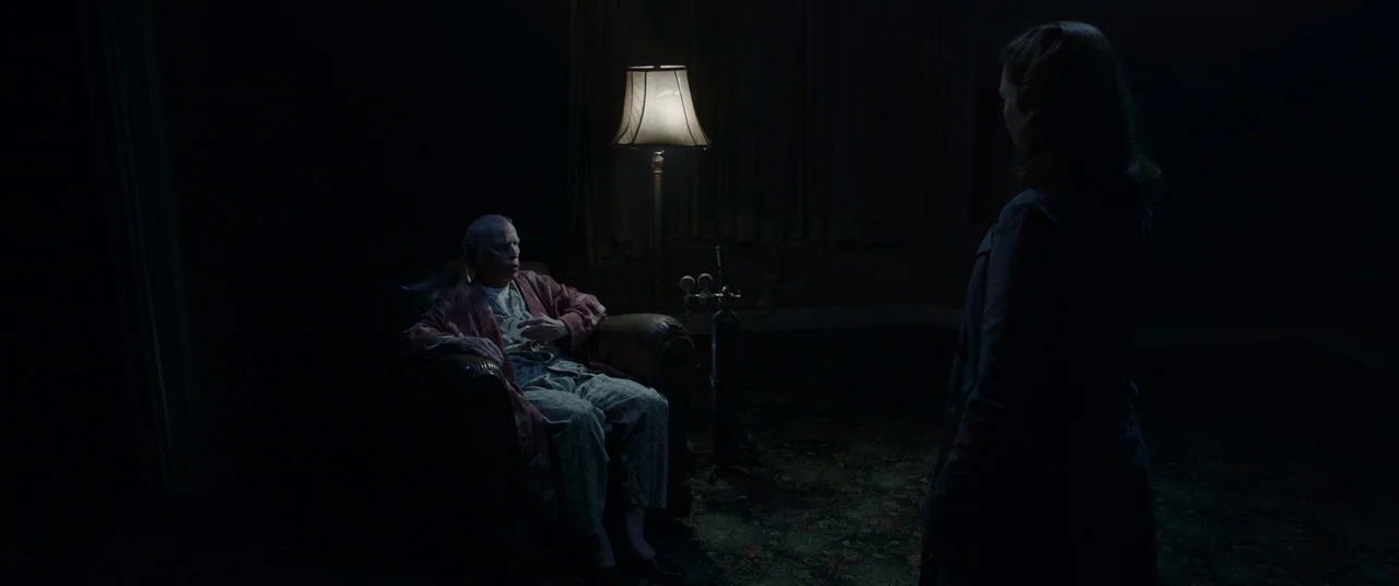 The Conjuring 2 (2016) movie still: wide — A dimly lit room with a patterned rug and dark furniture in deep shadow. An elderly man in pajamas sits…