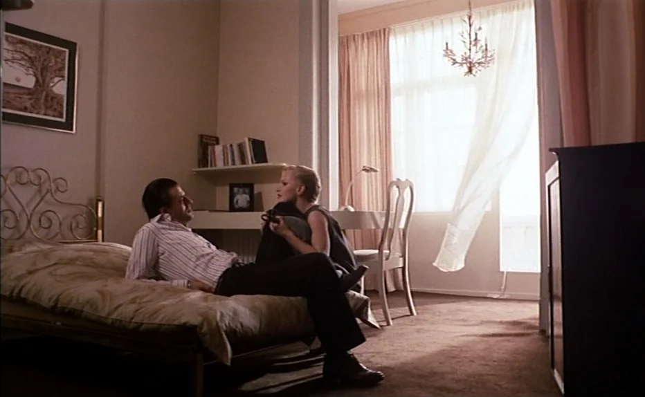 The Fourth Man (1983) movie still: wide — A hotel room features a bed, a desk, a chair, and sheer window curtains. A man sits on the edge of the bed…