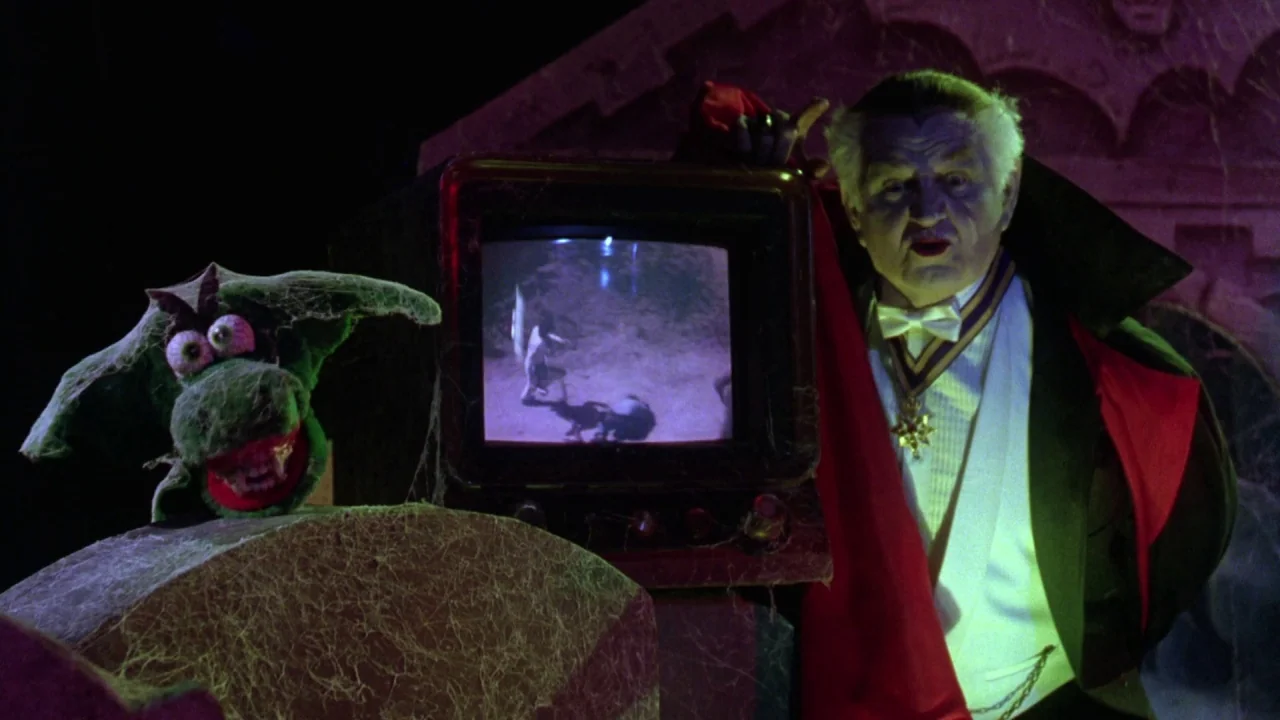 Gremlins 2: The New Batch movie still: medium — An elderly man dressed as a vampire stands next to an old television set. A small green puppet with…