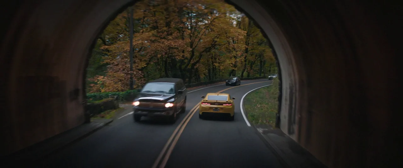 Pig (2021) movie still: wide — A dark curved tunnel exit frames a two-lane road flanked by autumn trees. A yellow sports car and a black SUV drive…
