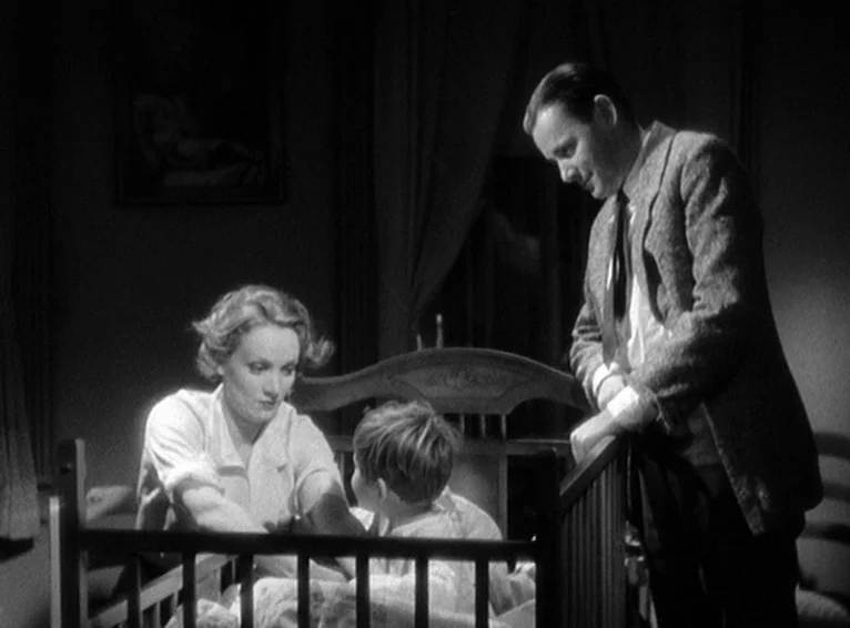 Blonde Venus (1932) movie still: medium — A dimly lit room contains a wooden crib centered in the foreground. A man and a woman stand by the railing…
