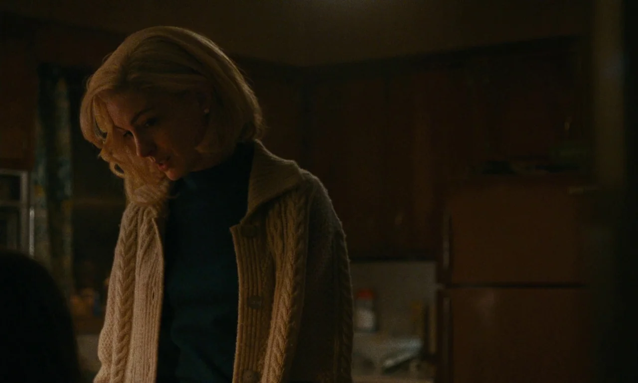 Eileen (2023) movie still: medium — A dimly lit room with dark wooden cabinetry visible in the soft-focus background. A woman in a thick cable-knit…