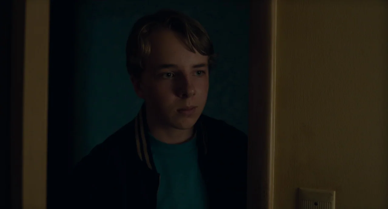 Wildlife (2018) movie still: closeup — A teenager stands in a dark doorway framed by a dimly lit yellow wall. The boy looks directly ahead with his…